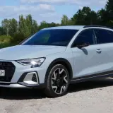 Jasnoszary Audi A3 Allstreet z czarnymi dodatkami stoi na żwirowej drodze nad jeziorem.