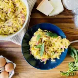 Un plat de pâtes à la carbonara crémeuses, garni de basilic frais. Des ingrédients comme des œufs, du fromage et de la farine sont visibles à proximité.