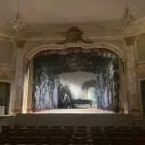 En ensam person vid ett piano på en scen med en skogsmålning. En vacker salong med kristallkronor, som påminner om **confidencen Ulriksdal**.
