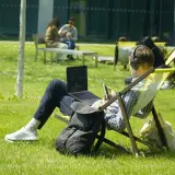 Mann mit Kopfhörern und Laptop im Freien, genießt sein Fernstudium im Ausland auf einer Liege im Sonnenschein.