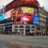 Tętniący życiem Piccadilly Circus w Londynie z neonowymi reklamami, w tym Samsung, McDonald's i Hyundai. Tłum ludzi na chodniku.