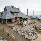 Drewniana chatka Puchatka w górach, z tarasem i kamiennym zboczem. W tle góry i niebo.