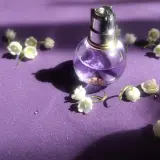 Szklana buteleczka perfum z fioletowym płynem i konwalią na fioletowym tle.