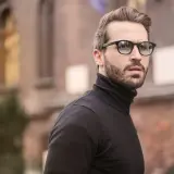 Hombre con barba y gafas, luciendo un jersey negro de cuello alto. Un estilo moderno que realza los rasgos, ideal para cortes para cara redonda hombre.