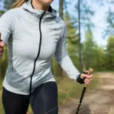 Kobieta w szarej kurtce i czarnych legginsach uprawia north walking na leśnej ścieżce, ciesząc się aktywnością na świeżym powietrzu.