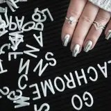 A hand with silver metallic nails and rings rests on a black surface scattered with white letters.
