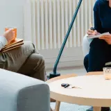 Dwie osoby siedzą w pokojach, jedna pije kawę, druga pisze.