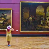 Kobieta w żółtej sukience i różowym kapeluszu podziwia obrazy w Luwrze. Co to za muzeum? To słynny paryski Luwr!