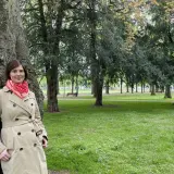 Kristina Hartenbach lehnt an einem Baum im Park. Sie trägt einen beigen Trenchcoat und einen roten Schal.