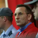 Słowik gangster w czerwonej marynarce, otoczony przez ochroniarzy w niebieskich koszulach i czapkach.