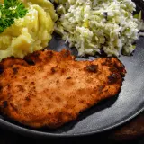 Kotlet schabowy z ziemniakami i surówką na ciemnym talerzu.