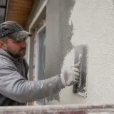 Budowlaniec nakłada tynk na elewację domu, używając kielni. Wybór odpowiedniego tynku na elewację jest kluczowy dla trwałości i wyglądu budynku.