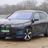 Ciemnoszary BMW iX z niebieskimi akcentami stoi nad rzeką, z mostem w tle.