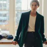 Elegancka kobieta w ciemnozielonej marynarce z aksamitu, stojąca przy drewnianym stole. Na stole czapka, szalik i pasek.