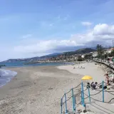 Plage de sable à Santo Stefano al Mare, avec des gens profitant du soleil et de la mer. La ville s'étend le long de la côte, bordée de collines verdoyantes.