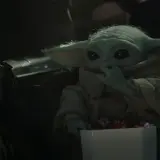 Dziecko z mandalorian film, w brązowym płaszczu, je słodycze z białego pudełka. Obok widać fragment zbroi Mandalorianina.