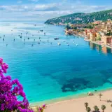 Blick auf einen malerischen Strand in Frankreich mit türkisfarbenem Wasser, Segelbooten und einer Küstenstadt, umrahmt von leuchtend lila Bougainvillea.