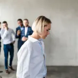 Smutna kobieta odwrócona tyłem, podczas gdy trzej mężczyźni za nią szepczą i wskazują na nią. To przykład mobbingu w pracy.