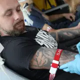 Mężczyzna z tatuażem na ramieniu oddaje krew. Czerwona opaska uciskowa i igła są widoczne.