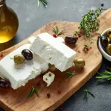Kostki sera feta, oliwki, rozmaryn i oliwa z oliwek na drewnianej desce. Idealne składniki na grecką sałatkę.