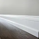 Biały, ozdobny cokół tworzy elegancką linię między ciemną, drewnianą podłogą a jasną ścianą.