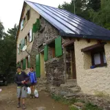 Llegada al refugio de Vallferrera. Dos jóvenes con mochilas y un hombre sentado revisando un mapa esperan en la entrada de piedra y estuco.
