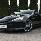 Czarny Aston Martin DB9 lśni na tle zielonych żywotników. Elegancki, sportowy samochód z charakterystycznym grillem i felgami.
