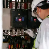 Elektryk w białym kasku i masce zabezpieczającej oznacza śruby przy głównym bezpieczniku prądu.
