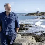 Ein Mann in blauer Kleidung steht am Meer. Die bannalec reihenfolge der Wellen spiegelt sich in seiner entspannten Haltung wider.