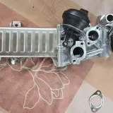 VW AGR-Kühler mit schwarzem Stellmotor und gelber Kappe auf gemustertem Untergrund.
