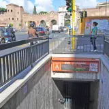 Ingresso metro San Giovanni, collegamento metro A. Traffico intenso con auto e scooter, sullo sfondo le antiche mura romane.