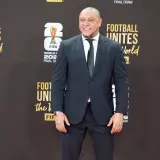 Roberto Carlos żyje i wita fanów na czerwonym dywanie, ubrany w elegancki garnitur. Uśmiecha się szeroko, a w tle widać logo FIFA i napisy związane z Mistrzostwami Świata.