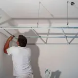 Mężczyzna w białej koszulce montuje metalowy stelaż pod sufity podwieszane. Widać przewody elektryczne w ścianie.
