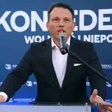 Mężczyzna w garniturze przemawia do mikrofonu na tle baneru Konfederacji. Dyskutuje, dlaczego KO chce odebrać dotacje partii opozycyjnej poprzez nowelizację kodeksu wyborczego.