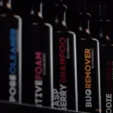 Rząd czarnych butelek z etykietami: "POSECLEANER", "CTIVEFOAM", "RASPBERRY SHAMPOO", "BUGREMOVER", "ODIE WHEELS", "ASSCLEANER". To są kosmetyki do samochodu.