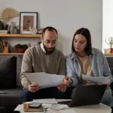 Pareja revisa documentos y laptop, debatiendo qué tasadora tasa más alto para su hipoteca.