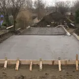 Plac budowy z wylana płytą betonową, przygotowywaną pod fundamenty. Widać zbrojenie i szalunki, wskazujące na różne klasy ekspozycji betonu.