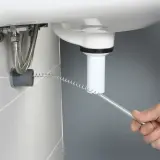 Ręka wprowadza sprężynę do rury pod umywalką, pokazując, jak udrożnić rury.