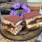 Dwa kawałki tiramisu, idealne ciasto do kawy, obok słoika z ziarnami kawy i fioletowych kwiatów.