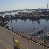 Widok z kamery portowej we Władysławowie. Wiele łodzi rybackich i jachtów zacumowanych w porcie, spokojna woda odbija niebo.