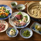 A spread of Japanese side dishes, including sashimi, edamame, potato salad, and a vegetable stir-fry, complements a pot of rice with mushrooms.
