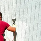Uomo con maglietta rossa e occhiali da sole sta pitturando casa a spruzzo con una pistola.