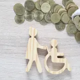 Drewniane figurki osoby pomagającej osobie na wózku inwalidzkim i monety. Czy zasiłek pielęgnacyjny wlicza się do dochodu?