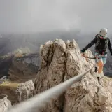 Kobieta na via ferrata polska pokonuje skalną grań, w tle górzysty krajobraz i kręta droga.