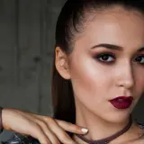 Kobieta z mocnym makijażem wieczorowym, ciemnymi ustami i oczami, w brązowej opasce na nadgarstku.