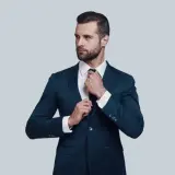 Elegancki mężczyzna w granatowym garniturze i białej koszuli poprawia krawat. Idealna stylizacja, gdy zastanawiasz się, jaka koszula do granatowego garnituru.