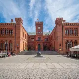 Rynek Kołobrzeg: ceglany ratusz z zegarem i flagą, przed nim kamienna rzeźba i brukowany plac.