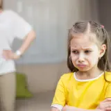 Smutna dziewczynka w żółtej bluzce, z matką w tle.