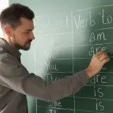 Nauczyciel pisze na tablicy odmianę czasownika "to be". Nauka angielskiego na poziomie C1 to cel wielu uczniów.