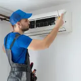 Technik w niebieskiej czapce i szarym ogrodniczkach naprawia klimatyzator śrubokrętem.